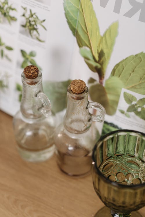 Free Glass bottles with cork stoppers beside a green goblet on a wooden surface with herb illustrations. Stock Photo